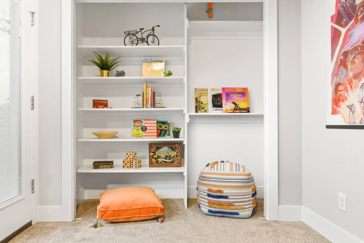 A playful, cozy nook with shelves filled with books and games, a colorful pouf, an orange pillow, and a decorative bicycle, all arranged in a small, inviting corner. The mood is lighthearted and creative, encouraging both relaxation and playful activities.