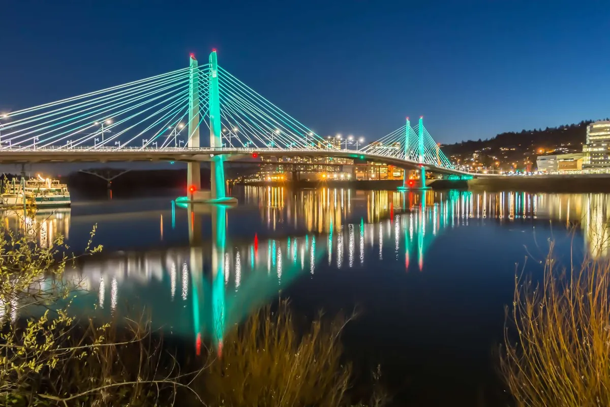 A dramatic nighttime cityscape showing an illuminated bridge reflected in the river below, set against a backdrop of sparkling city lights. The image captures the energy of Portland's urban environment and its lively riverfront.