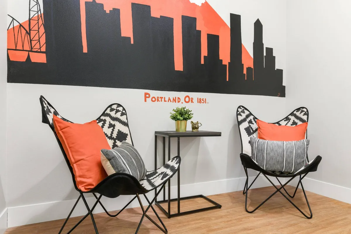 A whimsical seating nook adorned with a silhouette mural of the Portland skyline, colorful accent chairs, and a small side table with a potted plant. The artistic wall and cozy seating make it an ideal backdrop for selfies or quiet reflection.