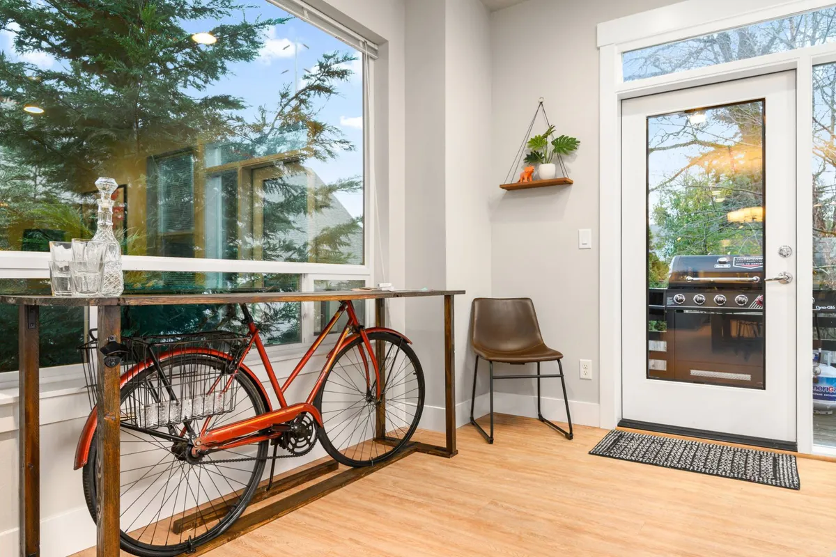 This image features a casual dining area with a bicycle visible near the table, glassware set out, and a doorway leading to an outdoor space. Natural light streams in, and the setup hints at effortless transitions between indoor relaxation and venturing out for work or school.