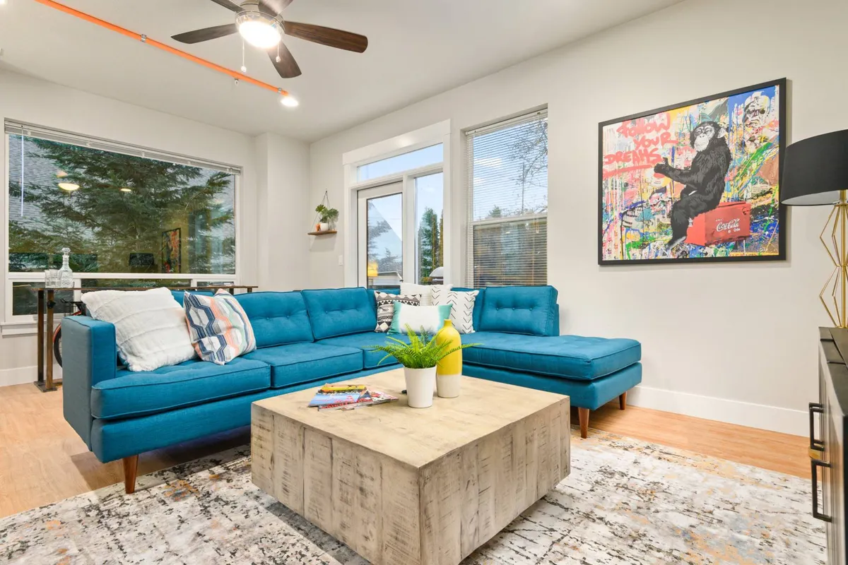 A vibrant, sunlit living room featuring a striking blue sectional sofa, contemporary artwork from local artists on the wall, a sleek coffee table, and large windows that bathe the space in natural light. The arrangement encourages conversation and relaxation.