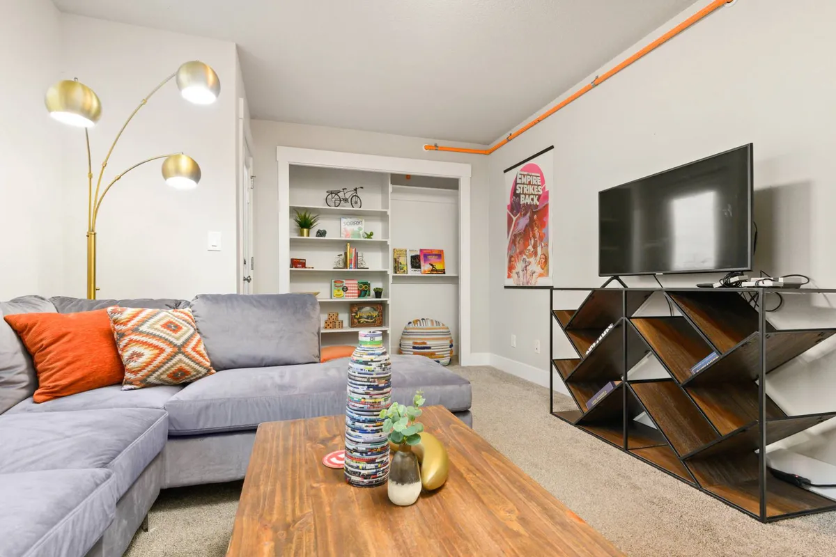 A cozy family lounge featuring a sofa with colorful cushions, a wooden coffee table, a bookshelf with homey decor, and a TV unit with a large TV. The space feels cheerful and welcoming, perfect for relaxing, playing games, or movie nights.
