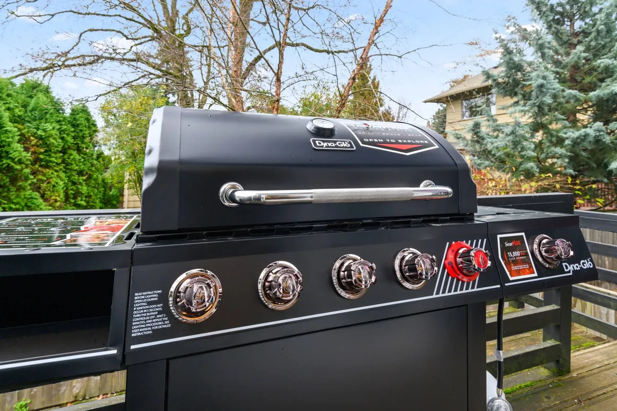 An outdoor backyard deck area featuring a grill, seating, and abundant greenery, ready for family barbecues or friendly gatherings. The space is bright and cheerful, ideal for outdoor relaxation and socializing.
