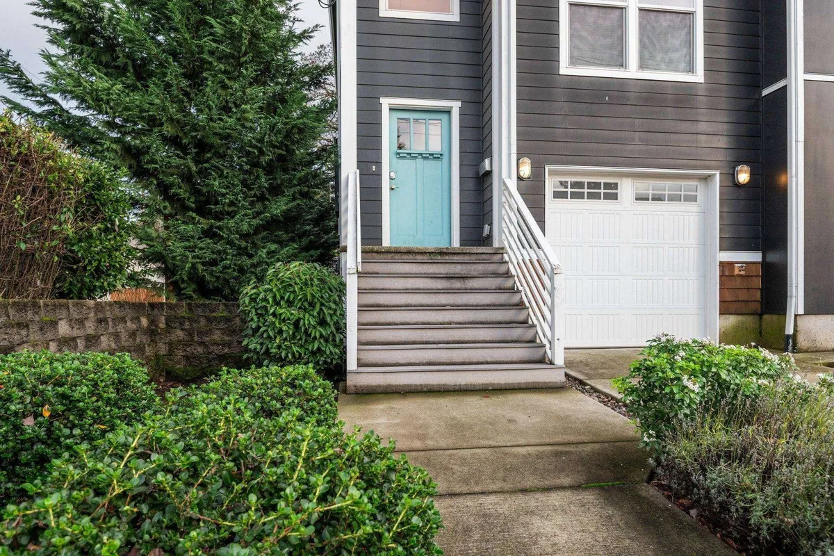 A bright exterior shot featuring a light blue front door, inviting stairs, and lush green shrubs lining the entrance. The garage door and porch area are also visible, establishing a sense of arrival and neighborhood charm.