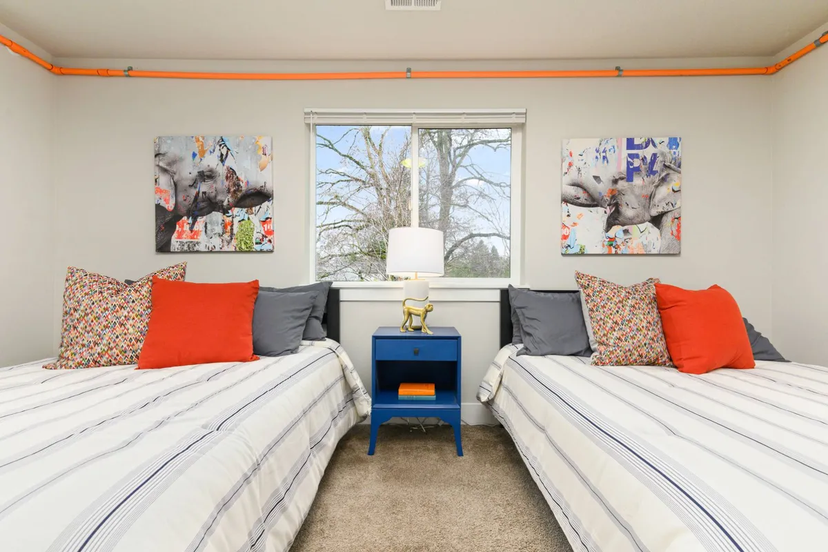 This image features two twin beds with colorful pillows, bright wall art, and a bedside lamp. Sunlight streams in, giving the room a cheery, inviting feel. The arrangement suggests flexible accommodations for siblings, friends, or extra guests.