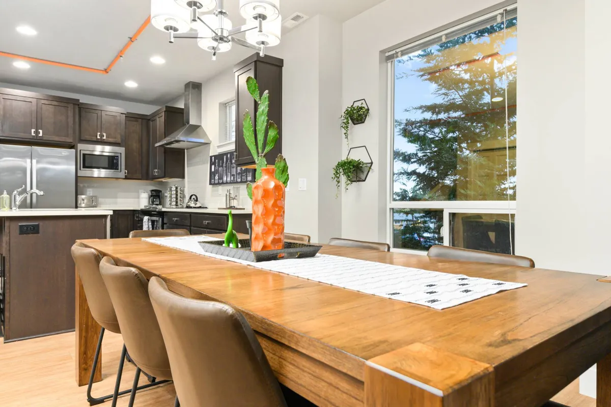 A warmly lit dining area with a spacious dining table, elegant chandelier, a vase with fresh greenery, and cabinetry that hints at the connected kitchen. The atmosphere invites group meals, celebrations, and intimate gatherings.