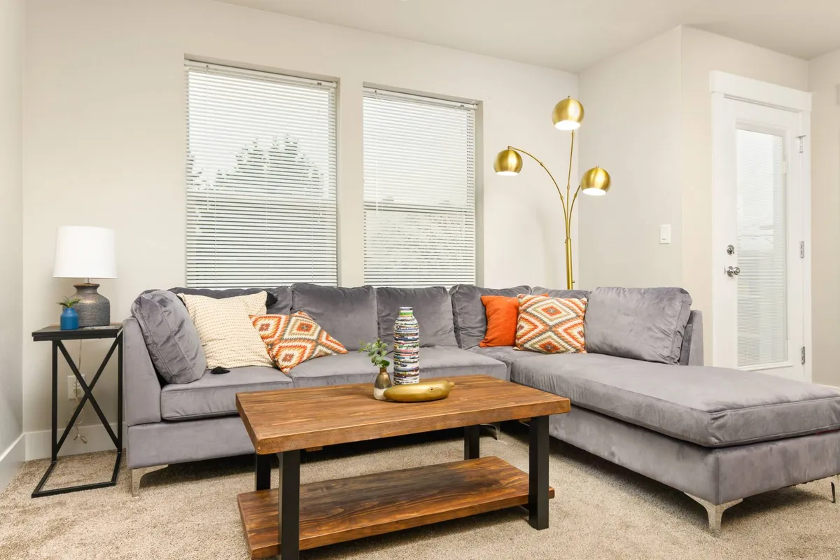 A bright, welcoming living room with a grey sectional sofa, decorative cushions, a wooden coffee table, and large windows that let in natural light. The room feels open, comfortable, and ideal for gatherings or quiet moments.