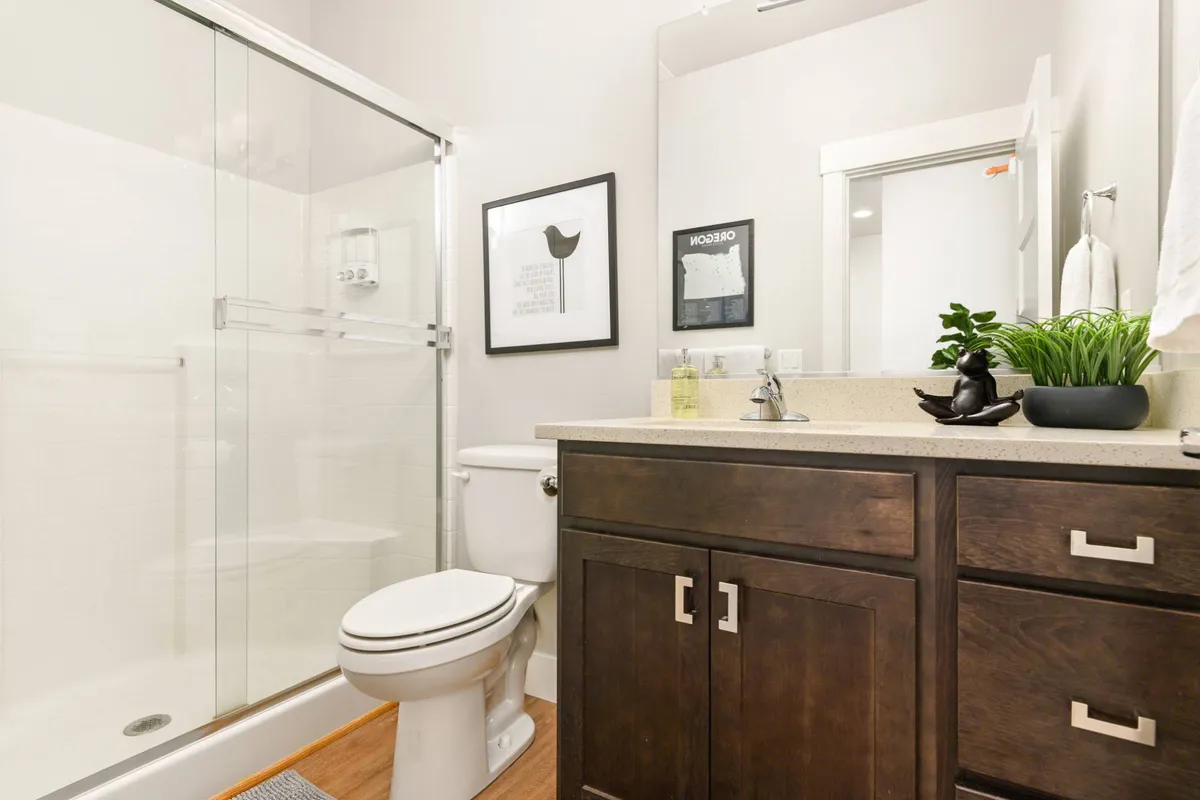 A serene bathroom with a glass shower stall, dark wood vanity, framed artwork, and a touch of greenery. The space feels inviting and private, ideal for relaxing or preparing for the day ahead.