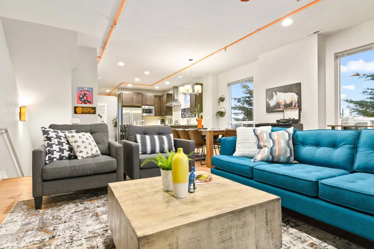 A modern, sunlit living space featuring a plush blue sofa, warm wooden coffee table, vibrant local artwork on the wall, and an inviting dining area in the background—perfect for gathering with friends and family.