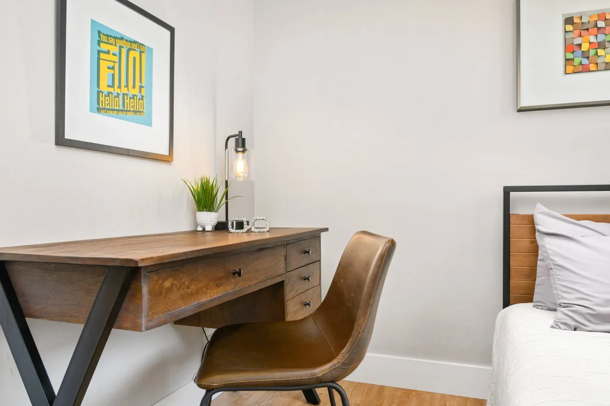 A cozy, sunlit workspace featuring a wooden desk and leather chair, accented with framed artwork, a table lamp, and a potted plant. The mood is peaceful, conducive to focused work or study.